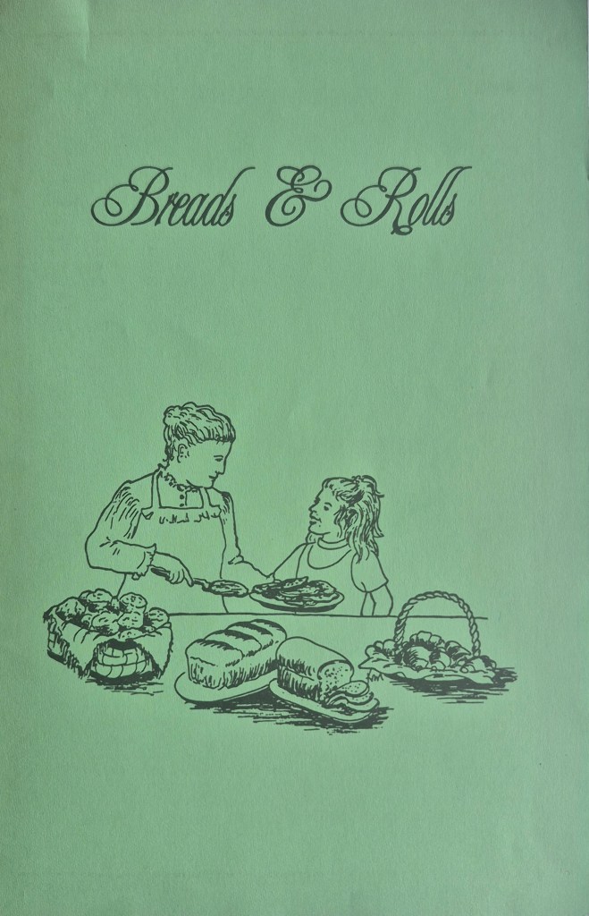 Breads and rolls artwork. An older woman in an apron hands over a plat of bread slices to a younger woman. There are loaves on the table in front of them. By Kathy McGill.